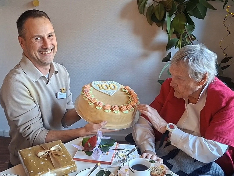 Martin Braungardt überreicht an einem Kaffetisch sitzend eine Torte mit einer verzierten 100