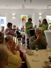 Zwei Pflegekräfte in grüner Arbeitskleidung singen und klatschen im voll besetzten und bunt geschmückten Saal bei der Wiedereröffnung der Tagespflegestätte für ältere Menschen im Immanuel Seniorenzentrum Kläre Weist in Petershagen