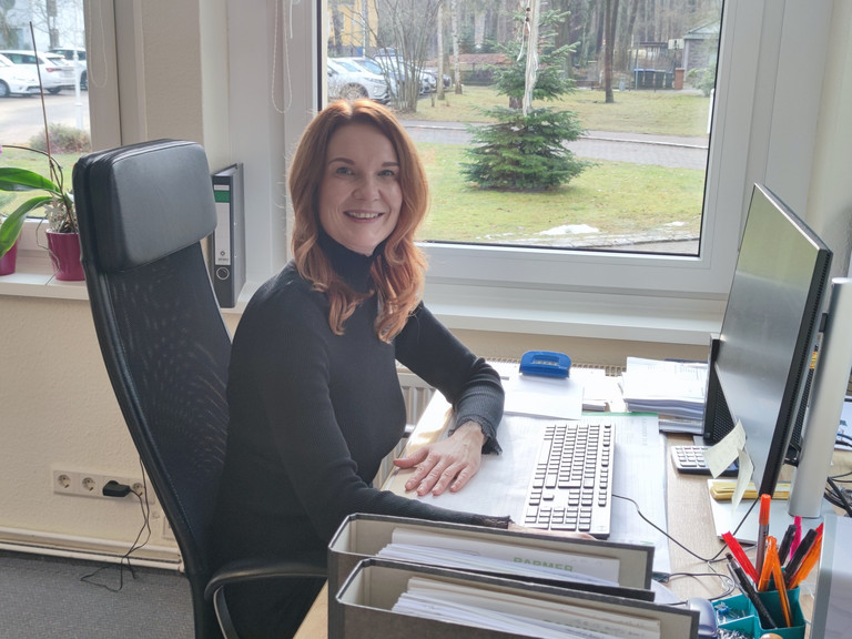 Umschülerin Christina blickt während ihres Praktikums im Immanuel Haus Ecktannen am Schreibtisch mit Monitor sitzend lächelnd in die Kamera