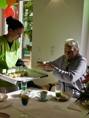 Eine Beschäftigte reicht einer am Tisch sitzenden Frau ein Tablett mit bunten Muffins bei der Wiedereröffnung der Tagespflegestätte für ältere Menschen im Immanuel Seniorenzentrum Kläre Weist in Petershagen