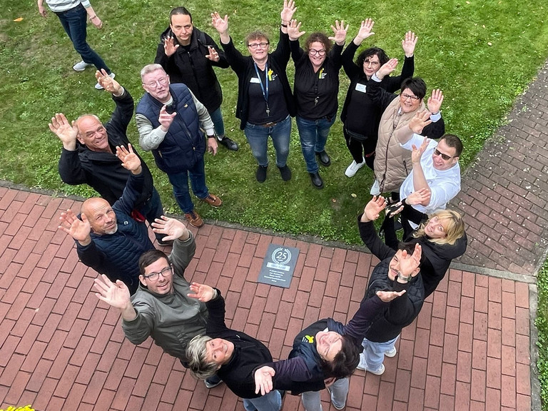Ein Team von Mitarbeitenden steht im Kreis und blickt nach oben die Kamera zum 25-jährigen Jubiläum des Immanuel Suchthilfeverbunds Guben mit besonderen Wohnformen für abhängigkeitskranke Menschen in Thüringen Ein Team von Mitarbeitenden steht im Kreis und blickt nach oben die Kamera zum 25-jährigen Jubiläum des Immanuel Suchthilfeverbunds Guben mit besonderen Wohnformen für abhängigkeitskranke Menschen in Thüringen