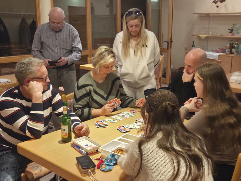 Mehrere Teammitglieder sitzen und stehen an einem Tisch beim Kartenspielen in der Immanuel Begegnungsstätte Ecktannen im Heilbad Waren beim gemeinsamen Spieleabend für Mitarbeitende der Seniorenpflegeeinrichtung mit Tagespflegeangebot Immanuel Haus Ecktannen.
